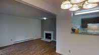 This single-floor residence offers a clean, straightforward layout designed for comfortable everyday living. The living area is a standout feature of the home, showcasing rich hardwood flooring that runs throughout and adds warmth to the space. A classic fireplace with a crisp white surround is set against a bold gray accent wall, serving as a natural focal point for the room and creating a cozy atmosphere. The ceiling features a textured finish, adding subtle visual interest overhead. A baseboard heating unit lines the wall, providing practical climate comfort throughout the cooler months. Adjacent to the living area, the dining space is illuminated by a decorative multi-globe ceiling light fixture that brings a touch of vintage charm to the interior. A granite or stone ledge is visible nearby, hinting at additional functional surfaces within the home. The overall color palette — soft whites, cool grays, and warm wood tones — creates a balanced, neutral aesthetic that is both modern and inviting. The open flow between the living and dining areas enhances the sense of space, making the home feel bright and airy despite its single-story footprint. This residence is well-suited for those seeking a practical yet stylish living environment.