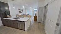 This 580-square-foot, one-bedroom, one-bathroom apartment at 13959 Hillcroft in Houston, TX features a modern, open-concept design with light wood-style flooring throughout. The combined kitchen and living area serves as the heart of the unit, where a sleek kitchen showcases dark wood cabinetry, white quartz countertops, stainless steel appliances—including a refrigerator, microwave, and range—and a peninsula island with pendant lighting and seating for four. The adjacent living room offers large windows that flood the space with natural light. The 140-square-foot primary bedroom includes multiple windows, a ceiling fan, and direct access to the bathroom. The well-appointed bathroom features a dark wood vanity with a white countertop, a large mirror, and contemporary light fixtures. A dedicated walk-in closet provides ample storage with multi-level wire shelving on three walls. The unit's clean, neutral palette and consistent modern finishes create a cohesive and functional living environment throughout.