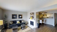 This single-story space in Arlington offers a well-appointed interior with a thoughtful layout that balances comfort and functionality. The living area serves as the central gathering space, highlighted by a white-mantled fireplace that anchors the room and adds a classic focal point. Hardwood-style flooring runs throughout, lending warmth and a clean, cohesive aesthetic to the space.

The living area is generously proportioned, providing ample room for seating arrangements, and is accented by neutral grey walls that create a versatile backdrop. Framed artwork adorns the walls, contributing to a polished, curated atmosphere. The space transitions openly into the dining area, which features a ceiling fan and white dining chairs, maintaining a bright and airy feel. A partial view into the kitchen suggests a connected, functional layout ideal for everyday living and entertaining.

Natural greenery in the form of potted plants adds life and texture to the space. The overall ambiance is modern yet inviting, with a neutral color palette punctuated by yellow and dark accent tones that add visual interest throughout.