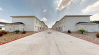 This multi-unit residential complex presents a clean, modern aesthetic defined by rows of two-story townhome-style buildings facing one another along a spacious central concrete drive. The buildings feature a neutral beige and white exterior finish with dark blue pitched rooflines, arched entryway accents, and individual unit doors — lending a cohesive yet welcoming character to the development. Each unit is equipped with multiple windows that allow for natural light, and ground-level entries provide direct outdoor access for residents.

The central drive is wide and well-marked with directional arrows, accommodating smooth traffic flow through the property. Red curbing borders the drive, clearly delineating the roadway from the surrounding landscape areas. The grounds are finished with gravel and rock landscaping accented by ornamental grasses, small palms, and drought-tolerant plantings, offering a low-maintenance yet visually appealing environment.

The main entrance opens directly from a public road, with mature trees visible along the perimeter providing a natural backdrop. Exterior utility connections and drainage infrastructure are thoughtfully integrated throughout the site. The overall layout and design reflect a practical, well-organized development suited for community living, with open skies and a spacious outdoor atmosphere defining the character of the entrance and shared areas.