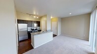 This single-floor space showcases an open-concept layout that integrates the kitchen and living area into one cohesive, functional environment. The kitchen is well-appointed with dark espresso cabinetry, stainless steel appliances including a refrigerator, microwave, and range, and granite-style countertops that add a polished finish. A peninsula with a built-in sink and additional counter space extends outward, creating a natural divide between the kitchen and the adjoining living area while also offering casual seating or prep space. Track lighting above the kitchen provides focused illumination, complementing the recessed lighting found throughout. The kitchen features rich dark hardwood-style flooring, which transitions into plush neutral-toned carpeting in the living area, offering a comfortable and welcoming atmosphere. The living space is open and generously proportioned, with neutral beige walls that lend a clean, versatile backdrop. A white interior door near the entry adds a classic touch. The overall design balances practicality with modern aesthetic details, making it a well-organized and inviting space.