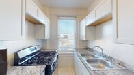 This single-floor unit offers a well-organized layout that makes efficient use of its space. Throughout the living and bedroom areas, warm hardwood flooring adds a classic character, complemented by crisp white walls and bold dark door trim that creates a striking contrast. The living space is generously lit by a row of windows that invite ample natural light, while a traditional radiator provides warmth during cooler months. The bedroom features the same rich hardwood floors, with dark-trimmed doorways and a mirrored closet door that adds depth to the room. The galley-style kitchen is thoughtfully designed with white cabinetry lining both walls, granite-style countertops, a gas range, a stainless steel refrigerator, a double-basin sink, and a window at the far end that brightens the cooking area with natural light. The tile flooring in the kitchen provides a clean and practical surface. The bathroom is neatly finished with white subway tile wainscoting, a white vanity with a mirror, and a tiled floor, creating a bright and functional space. A hallway connects the rooms efficiently, featuring the same hardwood flooring and dark trim detailing consistent throughout the unit. Overall, the space presents a cohesive and practical design with classic finishes.