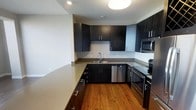 This contemporary single-floor space showcases a thoughtfully designed interior with a strong emphasis on modern aesthetics. The kitchen is a standout feature, outfitted with rich, dark flat-panel cabinetry that extends from floor to ceiling, offering generous storage capacity. Smooth, light-toned countertops provide a striking contrast against the dark cabinetry, creating a balanced and visually appealing palette. The kitchen is equipped with a full suite of stainless steel appliances, including a built-in microwave, dishwasher, and electric range, reflecting a commitment to both style and functionality. A deep undermount sink sits beneath a modern arc faucet, adding to the clean, streamlined look. Warm hardwood flooring runs throughout, lending the space an inviting and cohesive feel that complements the contemporary design. Recessed and flush-mount ceiling lighting ensures the space is well-illuminated, enhancing the open and airy atmosphere. The layout is efficiently arranged, with a peninsula-style counter extension that adds additional workspace and visual definition between areas. Overall, the space presents a refined, modern living environment with high-quality finishes and a cohesive design sensibility throughout.