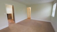 Located at 137 B S. Symington, this single-floor unit offers a straightforward and functional layout with a clean, neutral aesthetic throughout. The main living area is finished with wall-to-wall beige carpeting that provides a warm, comfortable feel underfoot, while the walls are painted in a soft, light green-white tone that creates a bright and airy atmosphere. White painted baseboards frame the perimeter of the room, adding a crisp, finished look to the space. A doorway visible from the main room leads to an adjacent area, which appears to connect to additional rooms, suggesting a practical and accessible floor plan. To one side, an open doorway reveals hardwood-style flooring, offering a transition between spaces and hinting at varied finishes throughout the unit. The overall design is simple and understated, making the space highly adaptable to a range of furnishing styles and personal aesthetics. The unit's single-floor configuration ensures easy navigation with no stairs, adding to its practicality and accessibility.