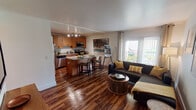 This single-floor residence offers a thoughtfully designed open-concept layout that seamlessly connects the kitchen, dining, and living areas. The kitchen is outfitted with warm honey-toned cabinetry, stainless steel appliances including a refrigerator, range, and microwave, and a granite-topped island that doubles as a casual dining space with bar seating — ideal for both cooking and entertaining. Track lighting overhead ensures the kitchen is well-lit at all times.

The living area is bright and welcoming, benefiting from a large sliding glass door that ushers in natural light and provides an outdoor view. Rich, dark hardwood-style flooring runs throughout the main living spaces, lending the home a warm and cohesive aesthetic.

The bedroom is a serene retreat, featuring the same hardwood-style flooring, a window with curtains that allow for flexible light control, and ample wall space. A closet provides convenient storage, and the room maintains a calm, neutral palette throughout.

The bathroom is clean and functional, featuring a full tub-and-shower combination, tile flooring, and a crisp, all-white finish that creates a fresh, airy feel. Overall, the residence balances practical design with a warm, contemporary ambiance.