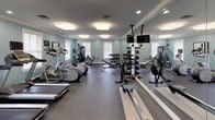Located at 5 Princeton Drive in Hooksett, New Hampshire, this fitness center is a comprehensively outfitted single-floor facility designed to support a wide range of workout routines. The space is finished with soft blue-gray walls, white crown molding, and a combination of flush and recessed lighting that keeps the environment bright and welcoming throughout.

The cardio area features multiple treadmills and elliptical machines arranged in a row, providing ample space for simultaneous use. A rowing machine and additional cardio equipment round out the aerobic offerings. For strength training, the facility includes a multi-station cable machine, a leg extension/curl machine, and an adjustable bench. A dedicated free weight section houses a full rack of dumbbells in graduated sizes, alongside a medicine ball rack stocked with a variety of weighted balls and a mounted flat-screen television for entertainment.

Large windows fitted with white blinds allow natural light to fill the room, while full-length mirrors along one wall visually expand the space and aid in form monitoring. Durable rubber flooring covers the entire workout area, providing cushioning and traction. The overall ambiance is clean, organized, and functional, making it a practical and inviting environment for fitness.