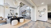 Located at 28 Seventh St NE in Rochester, Minnesota, this striking lobby is a masterclass in contemporary design. The space makes an immediate impression with its soaring double-height ceilings, which are anchored by cascading crystal chandeliers that draw the eye upward and fill the room with warm, ambient light. A dramatic floor-to-ceiling wall clad in large-format grey marble tile serves as the focal centerpiece, hosting a sleek linear fireplace and a mounted flat-screen display — a bold architectural statement that defines the lounge area.

The living room zone is thoughtfully appointed with tufted leather seating, a sculptural round coffee table, and a graphic area rug, creating a refined yet welcoming gathering space. Exposed industrial ductwork provides an urban contrast to the polished marble and crystal elements, adding depth to the design narrative.

The entry area features earthy stone-tile flooring, a substantial round pedestal table with floral arrangement, and a cluster of decorative circular mirrors that add visual interest to the crisp white walls. Large street-facing windows invite abundant natural light and offer views of the surrounding streetscape. A built-in reception counter with cabinetry completes the functional layout, making this lobby as practical as it is visually compelling.