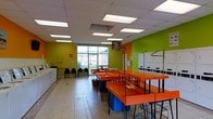 This single-floor laundromat is designed with both functionality and visual energy in mind. The interior is immediately distinguished by its bold lime green accent walls, which contrast with neutral tile flooring and a standard drop ceiling fitted with evenly spaced fluorescent panel lights, ensuring consistent, bright illumination throughout the space. A large window along the far wall allows natural light to filter in, adding openness to the room. The layout is practical and spacious, with a central row of vivid orange folding tables supported by black metal frames, providing ample surface area for sorting and folding laundry. Blue laundry baskets are positioned conveniently beneath the tables. Along the right side, a bank of white stacked washing and drying machines lines the wall, offering substantial capacity for patrons. Additional machines are visible along the left wall. A small seating area near the window features black chairs, giving customers a comfortable place to wait. The overall ambiance is lively and utilitarian, with high-contrast colors creating an energetic atmosphere while the well-organized layout prioritizes ease of use and efficient traffic flow throughout the facility.