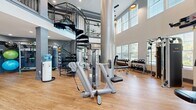 Located at 890 S Matlack St in West Chester, PA, this two-story fitness center offers a well-equipped and architecturally impressive workout environment spanning a generous total floor area across both levels. The main floor is defined by soaring double-height ceilings that create an open, airy atmosphere, complemented by large windows that flood the space with natural light and offer views of the surrounding greenery. Warm hardwood-style flooring runs throughout, contrasting elegantly with the bold dark-gray structural columns that anchor the layout. A wide array of strength and cardio equipment is thoughtfully arranged across the floor, including cable machines, weight benches, a full dumbbell and kettlebell rack, and various resistance training stations. A wall-mounted flat-screen TV and a water cooler add to the functionality of the main level. A striking black iron spiral staircase with diamond-plate treads serves as both a functional and aesthetic centerpiece, connecting the main floor to the upper level. The second floor continues the fitness theme with additional equipment, ceiling fans, pendant lighting, and an open railing that overlooks the main workout area below. Storage for exercise balls and accessories is neatly integrated into the lower level. The overall design balances industrial and modern elements, creating an inviting and highly functional fitness facility.