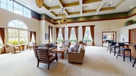 Located at 9000 Colby Dr in Fort Myers, Florida, this single-story community clubhouse offers a generously sized, well-appointed gathering space designed with classic Southern elegance. The grand entrance welcomes visitors through rich mahogany double doors framed by an arched transom window, decorative drapery, and intricate mosaic tile flooring. Stately white columns and a chandelier overhead set a refined tone from the moment of arrival. The expansive living room is a showpiece of architectural detail, boasting soaring coffered ceilings with bold wood trim, wide arched doorways, and plush neutral carpeting throughout. Large windows dressed in warm brown drapery flood the space with natural light while offering views of the lush Florida landscape, including palm trees and manicured greenery. A built-in bar and shelving area provides a functional yet stylish gathering point, complemented by seating areas arranged for conversation and relaxation. The game room is a standout feature, housing two full-size billiard tables beneath pendant lights, with floor-to-ceiling bookshelves lining the walls and arched windows bringing in additional natural light. Throughout the space, the design balances elegance and comfort, making it an inviting venue for community activities and social gatherings.