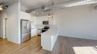 Located in the heart of Saint Paul, Minnesota, this single-floor unit blends industrial character with contemporary finishes throughout. The open-concept kitchen is a standout feature, showcasing full stainless steel appliances — including a French door refrigerator, electric range, microwave, and dishwasher — paired with crisp white cabinetry, a herringbone tile backsplash, and a generous white countertop island. Warm hardwood flooring anchors the kitchen and living areas, while pendant lighting and exposed metal ductwork overhead lend an authentic loft aesthetic. The carpeted main room offers a clean, neutral canvas, with high ceilings, recessed lighting, and visible ductwork continuing the industrial design language. A spacious walk-in closet provides generous wire-rack shelving for ample storage. Two bathrooms serve the space, each finished with classic black-and-white penny tile flooring and warm wood-toned vanities with white countertops. One bathroom features a soaking tub, while the other offers a walk-in shower stall, both equipped with grab bars and clean, functional fixtures. The overall ambiance is modern and move-in ready, balancing urban edge with everyday comfort in a well-connected downtown Saint Paul location.
