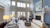 This impressive space makes an immediate statement with its dramatic two-story ceiling height, creating an expansive and airy atmosphere that defines the main living area. A sweeping wall of floor-to-ceiling windows and French doors floods the interior with natural light, offering views of the outdoor surroundings and seamlessly connecting the inside to the exterior. The living area is anchored by a classic white fireplace with a refined surround, set against a striking blue accent wall adorned with contemporary artwork — a bold design choice that adds depth and personality to the otherwise neutral palette. Soft grey tones dominate the furnishings and walls, complemented by warm hardwood flooring that runs throughout the space. A generous seating arrangement centered around a sculptural stone coffee table invites comfortable gathering, while accent chairs and carefully chosen décor contribute to a polished yet welcoming ambiance. A dining area positioned near the glass doors takes full advantage of the natural light and outdoor views. Crown molding and thoughtful architectural details elevate the overall design, making this a well-appointed space that balances elegance with everyday livability.