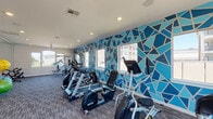 This fitness center in Houston, TX offers a comprehensive workout environment designed to meet a wide range of exercise needs. The space is defined by a bold, eye-catching geometric accent wall rendered in varying shades of blue, creating an energetic and modern atmosphere throughout. Large windows line the walls, flooding the interior with abundant natural light and providing views of the surrounding outdoor environment. The flooring consists of durable, patterned carpet tiles suited for high-traffic use. Recessed lighting and ceiling fans contribute to a comfortable and well-lit workout environment.

The cardio section is well-appointed with multiple ellipticals, stationary bikes, and treadmills, all equipped with personal display screens. For strength training, the facility includes a multi-station cable machine, a dedicated free weights area with a tiered dumbbell rack, and a large wall-to-wall mirror that enhances the sense of space while supporting proper form. Additional functional fitness equipment includes a heavy punching bag, stability and medicine balls in assorted sizes, and a wall-mounted television for entertainment. The open floor plan allows for easy circulation and flexible use of the space, making this fitness center a well-rounded facility for both cardio and strength-focused workouts.