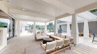 This single-story amenity facility is designed around an expansive outdoor lifestyle, seamlessly blending covered and open-air spaces for year-round enjoyment. The architecture features a classic white exterior with board-and-batten siding, crisp column detailing, and beadboard-clad ceilings that lend a polished, resort-like aesthetic throughout.

The covered patio areas serve as the heart of the space, offering generous seating arrangements with teak-framed sofas and lounge chairs upholstered in neutral tones. Recessed lighting and structural columns define each zone while maintaining open sightlines to the surrounding grounds. An outdoor fireplace paired with a wall-mounted television creates a focused gathering point, while black-framed glass doors connect the covered porch to the interior.

Beyond the covered areas, an open patio features a large sectional seating arrangement centered around a rectangular fire pit table, bordered by mature trees that provide natural privacy. Adjacent to this, a dedicated pool deck offers lounge chairs along a fenced swimming pool with open views of the landscaped surroundings.

The outdoor kitchen area is equipped with a stainless steel grill, granite countertops, a subway tile backsplash, and bold blue lower cabinetry accented by gray upper cabinets—combining functionality with sharp, contemporary style.