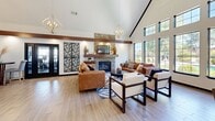 Located at 771 E Butler Rd in Mauldin, SC, this thoughtfully designed single-floor space offers an open-concept layout that seamlessly connects the living room, dining room, and kitchen into one cohesive, welcoming environment. Soaring vaulted ceilings with prominent wood beam accents create a dramatic sense of height and volume throughout, while light wood-toned flooring ties each area together with warmth and continuity. The living room serves as the centerpiece of the space, anchored by a striking stacked-stone fireplace with a wood mantel and floating shelves. Elegant chandelier lighting and large multi-pane windows flood the area with natural light and provide outdoor views, enhancing the bright, airy ambiance. A bold botanical metal wall panel adds an artistic focal point near the entryway. The dining area accommodates a large table beneath a gold candelabra-style chandelier, complemented by tall windows that further brighten the space. The kitchen features warm wood cabinetry, granite-style countertops, stainless steel appliances, and a peninsula with bar seating, offering both functionality and style. Recessed lighting and pendant fixtures complete the polished, modern aesthetic throughout this well-appointed space.