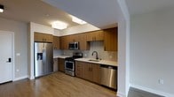 This single-floor space showcases a thoughtfully designed kitchen that serves as its centerpiece. The kitchen features warm wood-toned cabinetry extending from floor to ceiling, providing generous storage while lending a contemporary yet inviting aesthetic. A full suite of stainless steel appliances includes a French door refrigerator, gas range with oven, built-in microwave, and dishwasher, offering a fully equipped culinary environment. The countertops are finished in a clean, light-toned surface that complements the cabinetry, while a neutral grey subway tile backsplash adds texture and visual interest to the space. A deep single-basin sink with a modern matte black faucet is conveniently positioned beneath natural light. The flooring throughout is a light hardwood, contributing warmth and continuity across the space. Overhead, flush-mount ceiling fixtures provide consistent, well-distributed lighting. The open layout and clean sightlines create a sense of spaciousness, while the cohesive material palette—warm wood, stainless steel, and soft neutrals—gives the space a polished, modern character suited for everyday living.