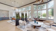 This sophisticated single-floor community clubhouse showcases a thoughtfully designed open layout bathed in natural light. Soaring ceilings with decorative crown molding create a grand sense of volume throughout, while expansive floor-to-ceiling windows along the main lounge area frame lush, mature tree-lined views and glimpses of a resort-style pool beyond.

The main lounge area is elegantly appointed with curved, upholstered accent chairs, a round marble-top coffee table, and patterned area rugs atop light hardwood flooring. Dramatic multi-arm chandeliers provide warm overhead illumination, complementing the bright, airy atmosphere cultivated by the abundant glazing. Tall, sculptural planters filled with ornamental grasses serve as stylish natural dividers between functional zones.

A dedicated dining area features a long rectangular table surrounded by cognac-toned leather chairs, set beneath contemporary abstract artwork and additional chandelier lighting — ideal for gatherings and meetings. French doors open directly to the outdoor courtyard, enhancing the seamless indoor-outdoor flow.

A well-equipped kitchenette includes sleek slate-blue cabinetry, white countertops, a matte black faucet, and a built-in wine refrigerator, all accented by a striking geometric mosaic tile backsplash. The entry vestibule offers a welcoming transition with tall windows and curated décor, completing this polished and functional community space.