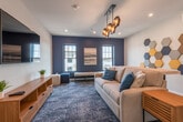 This thoughtfully designed single-floor lounge space combines contemporary style with comfort and functionality. The room is anchored by a large wall-mounted flat-screen TV set against a crisp white wall, complemented by a warm wood media console with drawers for storage. A generously sized sectional sofa in neutral tones, accented with teal throw pillows, sits atop a richly textured blue-toned area rug that adds depth and warmth to the space.

A striking deep navy accent wall serves as a bold focal point, framing two black-trimmed windows that flood the room with natural light and offer views of the surrounding neighborhood. Between the windows, a coastal landscape painting adds a subtle decorative touch.

Overhead, a geometric multi-pendant chandelier with a gold finish serves as a sculptural lighting centerpiece, drawing the eye upward and reinforcing the modern aesthetic. Recessed lighting throughout provides additional illumination.

One wall features a decorative arrangement of hexagonal acoustic panels in shades of yellow, navy, blue, and gray — a functional yet artistic element that enhances both the room's acoustics and visual appeal. A wall-mounted mini-split HVAC unit ensures year-round climate comfort.

A live-edge wood communal table paired with the sofa offers a versatile surface for casual gatherings. Glass-paneled double doors complete the space, connecting it seamlessly to adjacent areas.
