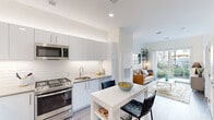 This well-designed single-floor residence offers a bright, contemporary living experience with a thoughtful open-concept layout. The kitchen is a standout feature, outfitted with sleek flat-panel cabinetry in soft white and grey tones, stainless steel appliances including a gas range, over-the-range microwave, and a French door refrigerator. A white tile backsplash adds a clean, modern touch, while light wood-look flooring flows throughout the main living areas. A compact dining island with bar seating creates a seamless transition between the kitchen and the living space. The living room is bathed in natural light courtesy of a sliding glass door that opens to a verdant outdoor area, offering a refreshing indoor-outdoor connection. A lean open shelving unit and a grid-style wall mirror contribute to the room's modern aesthetic. The bedroom is a serene retreat, featuring plush carpet, large windows framed by elegant draping curtains, and peaceful garden views that bring the outdoors in. The bathroom is finished in crisp white with a full soaking tub and shower combination. Throughout the space, high ceilings and recessed lighting enhance the overall sense of openness and airiness.