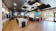 Located in Wilson, NC, this thoughtfully designed single-floor amenity facility offers a dynamic blend of social, recreational, and wellness spaces across a generously sized footprint. The main lounge area serves as the heart of the facility, featuring warm hardwood-style flooring, a dramatic coffered ceiling with bold dark framing and decorative inset panels, and an eclectic mix of pendant lighting. Exposed brick walls, teal accent shelving, and curated artwork create a vibrant, modern atmosphere. A central island with bar seating anchors the space, while multiple lounge seating arrangements invite relaxed socializing. Large windows and glass-panel doors flood the area with natural light and provide views of the outdoor surroundings. A cozy lounge nook features wall-mounted TVs, framed artwork, and intimate café-style seating. The fully equipped fitness center is outfitted with racks, free weights, cardio machines, and ceiling fans, all set against deep teal-painted walls with ample natural light from surrounding windows. Adjacent to the gym, a mirrored yoga and group fitness studio features bold patterned wallcovering, polished concrete flooring, and wall-mounted displays. A dedicated game room houses a pool table, large-screen TV, and casual seating, completing this well-appointed community space.