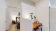This single-floor residence in Berkeley, CA offers a thoughtfully designed open layout with a clean, contemporary aesthetic throughout. Light wood-tone flooring and crisp white walls create a bright, cohesive atmosphere across all areas of the unit.

The living area is anchored by expansive floor-to-ceiling windows with bold black frames, flooding the space with natural light and offering open views of the surrounding Berkeley cityscape. A wall-mounted mini-split system provides efficient climate control. Recessed lighting complements the natural light throughout.

The kitchen is outfitted with high-gloss white cabinetry, light quartz countertops, a textured tile backsplash, and a full suite of stainless steel appliances, including a refrigerator, range, microwave, and dishwasher. A glass-top dining table with modern chairs fits neatly within the kitchen area.

A partial wall divider defines the sleeping area from a built-in workspace featuring a warm wood desk, offering functional separation without sacrificing openness. The bathroom includes a full soaking tub and shower combination with clean white finishes and light wood-look flooring, maintaining the unit's consistent modern style.