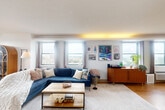 Located at 401 Cumberland Ave in the heart of Portland, Maine, this well-appointed one-bedroom, two-bathroom residence offers a thoughtfully designed open-concept layout with warm hardwood flooring throughout. The kitchen is a standout feature, showcasing sleek white cabinetry, a striking dark hexagonal tile backsplash, stainless steel appliances, and a tiled peninsula island that flows seamlessly into the main living area — ideal for both cooking and entertaining. Recessed lighting and open sightlines give the combined kitchen and living space an airy, connected feel. The expansive living room is bathed in natural light from a series of large windows framing panoramic views of the Portland skyline and surrounding greenery, with ambient cove lighting adding warmth to the atmosphere. The generously sized primary bedroom continues the view experience with floor-to-ceiling windows, built-in shelving, and ample closet space. A cozy secondary room with built-in shelves provides flexible living options. Both bathrooms are finished in crisp white tile, each featuring a full tub and shower combination. Additional conveniences include an in-unit laundry room and a private balcony, completing this well-rounded urban residence in one of Maine's most vibrant neighborhoods.