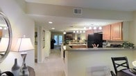 This single-floor residence showcases an open-concept layout that effortlessly connects the kitchen, dining, and living areas into one cohesive and functional living space. The kitchen is well-appointed with warm wood cabinetry, dark countertops, and a black refrigerator, complemented by a peninsula with bar seating that serves as a natural divider between the kitchen and the main living area. Pendant lighting above the peninsula adds a stylish focal point, while recessed lighting throughout ensures the space remains bright and inviting. The living area is carpeted in a soft neutral tone, contributing to the warm and comfortable ambiance of the home. A ceiling fan overhead adds both practicality and a classic touch. Framed artwork lines the hallway walls, lending a personalized and curated feel to the interior corridors. French doors at the far end of the living area invite natural light deep into the home and provide a seamless connection to the outdoors. The overall color palette of soft beiges and warm neutrals creates a cohesive, welcoming atmosphere throughout, making this a well-designed and comfortable single-level living space.