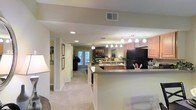 This single-floor residence showcases an open-concept layout that effortlessly connects the kitchen, dining, and living areas into one cohesive and functional living space. The kitchen is well-appointed with warm wood cabinetry, dark countertops, and a black refrigerator, complemented by a peninsula with bar seating that serves as a natural divider between the kitchen and the main living area. Pendant lighting above the peninsula adds a stylish focal point, while recessed lighting throughout ensures the space remains bright and inviting. The living area is carpeted in a soft neutral tone, contributing to the warm and comfortable ambiance of the home. A ceiling fan overhead adds both practicality and a classic touch. Framed artwork lines the hallway walls, lending a personalized and curated feel to the interior corridors. French doors at the far end of the living area invite natural light deep into the home and provide a seamless connection to the outdoors. The overall color palette of soft beiges and warm neutrals creates a cohesive, welcoming atmosphere throughout, making this a well-designed and comfortable single-level living space.
