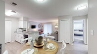 This single-floor residence is designed with an open-concept layout that seamlessly connects the main living areas. The galley-style kitchen is outfitted with white shaker cabinetry, granite-look countertops, black appliances including a refrigerator, range, and built-in microwave, and a double sink. Light wood-look flooring runs throughout the kitchen, while a peninsula counter separates it from the adjacent dining area, which accommodates a round wood dining table.

The dining space flows directly into the living room, where plush carpet continues underfoot. The living room offers a relaxed, functional layout with a wall-mounted television and a sliding glass door with vertical blinds that brings in natural light. Neutral gray walls carry throughout, contributing to a calm and cohesive aesthetic.

The bedroom is generously proportioned, featuring soft carpet, two closets providing ample storage, and multiple windows that allow abundant natural light to fill the room. The bathroom is clean and well-appointed with a white vanity, large mirror, and a shower-tub combination with a curtain. Overall, the space presents a practical and comfortable single-floor layout with consistent, neutral finishes throughout.