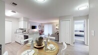 This single-floor residence is designed with an open-concept layout that seamlessly connects the main living areas. The galley-style kitchen is outfitted with white shaker cabinetry, granite-look countertops, black appliances including a refrigerator, range, and built-in microwave, and a double sink. Light wood-look flooring runs throughout the kitchen, while a peninsula counter separates it from the adjacent dining area, which accommodates a round wood dining table.

The dining space flows directly into the living room, where plush carpet continues underfoot. The living room offers a relaxed, functional layout with a wall-mounted television and a sliding glass door with vertical blinds that brings in natural light. Neutral gray walls carry throughout, contributing to a calm and cohesive aesthetic.

The bedroom is generously proportioned, featuring soft carpet, two closets providing ample storage, and multiple windows that allow abundant natural light to fill the room. The bathroom is clean and well-appointed with a white vanity, large mirror, and a shower-tub combination with a curtain. Overall, the space presents a practical and comfortable single-floor layout with consistent, neutral finishes throughout.