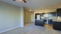 This single-floor space offers an open-concept layout that seamlessly connects the living area to the kitchen. The living area is finished with neutral-toned carpet and painted walls, contributing to a clean, understated aesthetic. A ceiling fan overhead provides both functionality and a subtle design element.

The kitchen is well-appointed with dark espresso cabinetry that contrasts with lighter countertops and a central island that doubles as a breakfast bar. Stainless steel appliances, including a refrigerator, stove, and microwave, add a modern touch. Track lighting above the kitchen provides focused, practical illumination. The kitchen flooring transitions from the carpeted living area, further distinguishing the two zones.

A hallway visible in the background leads to additional rooms, suggesting a practical and efficient floor plan. The neutral color palette throughout—soft greige walls, light ceilings, and white trim—creates a cohesive and inviting atmosphere. The space is designed with everyday functionality in mind, balancing open communal areas with defined private spaces accessible via the connecting corridor.