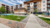 Located at 186 E Future Way in Draper, Utah, this outdoor community amenity space offers a well-rounded array of recreational facilities set against the stunning backdrop of the Wasatch Mountains. The grounds are thoughtfully designed to encourage an active lifestyle, featuring multiple sport courts — including a full basketball court and two tennis/pickleball courts — enclosed by chain-link fencing for safety and organization. A vibrant playground area with colorful play structures, slides, and benches sits along a winding concrete pathway, providing a welcoming space for younger residents. Manicured lawns, ornamental gravel landscaping, and young trees add visual appeal and a sense of order throughout the grounds. The surrounding multi-story residential buildings are constructed with a blend of stucco, stone accents, and warm wood-tone paneling, complementing the natural high-desert setting. A gated entry point connects the amenity area to the residential buildings, while a generously sized paved parking lot with clearly marked stalls ensures convenient access. Open skies and the rolling sagebrush hillsides beyond the property create a picturesque natural setting, lending the entire space an expansive, outdoor-oriented ambiance year-round.
