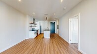 This single-floor unit showcases a clean, contemporary design with warm hardwood-style flooring running throughout the entire space. The open-concept layout centers around a well-appointed kitchen featuring a large central island topped with dark granite countertops and a matte black sink and faucet. White cabinetry, a stainless steel range with a wall-mounted hood vent, open wooden shelving, and a subway tile backsplash complete the kitchen. Track lighting overhead adds a modern, functional touch.

The living area extends naturally from the kitchen and is bright, featuring French doors that open to a balcony, allowing natural light to pour in. Bedrooms are generously proportioned, with one offering a wide window with neighborhood views and a ceiling fan. A walk-in closet equipped with adjustable wire shelving and hanging rods provides ample storage.

The bathroom maintains the same modern aesthetic, with a white vanity, dark granite countertop, matte black fixtures, and a full tub-shower combination. Crisp white walls and recessed lighting are consistent throughout, contributing to the unit's fresh, cohesive ambiance.