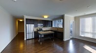 This single-floor residence showcases a thoughtfully designed open-concept layout where the kitchen flows seamlessly into the living area. The kitchen is a standout feature, outfitted with rich, dark espresso cabinetry that extends to the upper walls, providing generous storage space. Stainless steel appliances, including a full-size refrigerator, microwave, and range, lend a modern, polished aesthetic. A neutral tile backsplash complements the darker tones of the cabinetry, while a compact kitchen island with a granite-style surface and lower shelf adds both workspace and storage versatility. The sink is positioned beneath a window fitted with horizontal blinds, allowing natural light to filter into the space. Deep-toned hardwood flooring runs throughout, adding warmth and visual continuity between the kitchen and the adjoining living area. Crown molding trims the ceilings, contributing an element of classic architectural detail. A door near the kitchen suggests access to an outdoor area, potentially a balcony or patio, allowing for additional natural light. The overall ambiance is modern and functional, balancing dark finishes with open space.