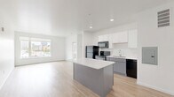 Located at 195 E Main St in the heart of downtown Columbus, Ohio, this one-bedroom, one-bathroom unit offers a clean, modern design across a single floor. Light-toned hardwood flooring runs throughout, creating a bright and cohesive aesthetic complemented by crisp white walls and neutral finishes.

The combined kitchen and living area forms the core of the unit. The kitchen is equipped with black appliances — including a refrigerator, range, and over-the-range microwave — white upper cabinetry, and a stainless steel sink. A central island with a white countertop and gray base provides additional prep space and casual seating, while recessed lighting overhead enhances functionality.

The living area is bathed in natural light through large windows that frame views of the Columbus skyline and surrounding urban streetscape, bringing the energy of the city directly into the space. The bedroom, though compact, features the same light hardwood flooring and a large window offering additional city views.

The bathroom maintains the unit's clean aesthetic with light wood-look flooring, a white vanity with ample drawer storage, brushed nickel hardware, and a well-lit mirror. The overall layout is efficient and thoughtfully designed, making strong use of the available footprint for comfortable urban living.
