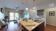 Located at 3600 N Westgate Dr in Weslaco, Texas, this single-story unit offers a thoughtfully designed open-concept layout finished with rich hardwood flooring throughout. The kitchen serves as a central hub, featuring dark espresso cabinetry that extends to the ceiling, a full suite of black appliances including a refrigerator, electric range, and microwave, and a peninsula island with a double-basin sink and light quartz-style countertops — ideal for both meal preparation and casual dining. Pendant lighting overhead adds a warm, inviting glow to the space. The kitchen flows seamlessly into the dining and living areas, where French doors open to a private balcony, filling the room with natural light and offering outdoor access. A ceiling fan completes the comfortable living space. The unit includes multiple bedrooms, each with hardwood flooring, ceiling fans, and generous natural light through large windows with blinds. One bedroom features notably elevated ceilings and direct access to an en suite bathroom. The bathrooms are finished with dark vanity cabinetry, light countertops, and wood-look flooring. A long hallway connects the various living spaces, contributing to the unit's efficient and well-organized single-floor layout.