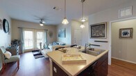 Located at 3600 N Westgate Dr in Weslaco, Texas, this single-story unit offers a thoughtfully designed open-concept layout finished with rich hardwood flooring throughout. The kitchen serves as a central hub, featuring dark espresso cabinetry that extends to the ceiling, a full suite of black appliances including a refrigerator, electric range, and microwave, and a peninsula island with a double-basin sink and light quartz-style countertops — ideal for both meal preparation and casual dining. Pendant lighting overhead adds a warm, inviting glow to the space. The kitchen flows seamlessly into the dining and living areas, where French doors open to a private balcony, filling the room with natural light and offering outdoor access. A ceiling fan completes the comfortable living space. The unit includes multiple bedrooms, each with hardwood flooring, ceiling fans, and generous natural light through large windows with blinds. One bedroom features notably elevated ceilings and direct access to an en suite bathroom. The bathrooms are finished with dark vanity cabinetry, light countertops, and wood-look flooring. A long hallway connects the various living spaces, contributing to the unit's efficient and well-organized single-floor layout.