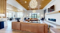 Located at 4275 Valley St in Enola, PA, this sophisticated single-floor community clubhouse offers a generously sized open layout designed for both comfort and style. Guests are welcomed by an inviting entry foyer accented with striking globe chandeliers and light hardwood flooring that flows throughout the entire space.

The central lounge area serves as the heart of the clubhouse, featuring a dramatic white brick fireplace with a natural wood mantel and wall-mounted television. Plush leather sectional seating surrounds the fireplace, while expansive black-framed windows and glass doors flood the space with abundant natural light and offer views of the surrounding community.

The full kitchen is outfitted with deep navy shaker-style cabinetry, crisp white countertops, a large center island with bar seating, stainless steel appliances, and a beadboard tile backsplash. Oversized globe pendant lights illuminate the space overhead.

Adjacent dining and lounge nooks provide flexible seating arrangements, including high-top bar tables and cozy accent chairs positioned beneath floor-to-ceiling windows. Throughout the space, warm wood-paneled tray ceilings add architectural depth and a refined, contemporary character to this well-appointed community gathering space.