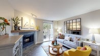 This thoughtfully designed single-floor residence offers a cohesive layout that balances comfort and functionality across its key living spaces. The living room serves as the heart of the home, anchored by a tile-surround fireplace with a rustic wood mantle and complemented by wood-look flooring, track lighting, and a large grid-panel mirror accent wall. Sliding glass doors flood the space with natural light and open directly onto a private outdoor deck, extending the living area outdoors. Adjacent to the living room, a compact dining nook provides an intimate setting for meals, accented by warm neutral tones and tasteful wall art. The galley-style kitchen is well-equipped with stainless steel appliances, granite-look countertops, crisp white cabinetry, and wood-look flooring that carries through from the main living areas. In-unit laundry is conveniently accessible from the kitchen corridor. The two bedrooms feature plush carpeting, ample natural light through large windows, and generous closet space, including a mirrored sliding-door closet in the primary bedroom. The full bathroom is finished with a granite-look vanity countertop, white cabinetry, wood-look flooring, and a shower-tub combination. Throughout the residence, a neutral color palette creates a warm, inviting ambiance.