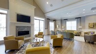 Located at 199 Cedar Park Boulevard in Northampton County, Pennsylvania, this single-floor space offers a refined and welcoming environment designed for comfort and style. The interior is anchored by a stunning lounge area that immediately draws attention with its soaring vaulted ceiling, accented by crisp white trim and recessed lighting that creates a bright, airy atmosphere. A floor-to-ceiling marble fireplace serves as a dramatic focal point, framed by a classic white mantel that adds an elegant, traditional touch. The hardwood flooring throughout lends warmth and continuity to the space, complementing the soft gray wall tones and white crown molding. Seating is thoughtfully arranged around a mirrored cocktail table, with plush chartreuse armchairs and muted sage sofas creating inviting conversation areas. Large windows dressed with patterned drapery flood the interior with natural light and offer views of the surrounding exterior. A wall-mounted flat-screen television is seamlessly integrated into the lounge area, enhancing the space's functionality. The overall ambiance strikes a balance between upscale residential comfort and polished communal design, making it an ideal gathering place for residents and guests alike.