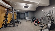 Located at 5000 Euclid Ave in Cleveland's vibrant arts corridor, this single-floor amenity space offers a well-appointed combination of fitness and social areas spanning a generous footprint. The fitness center is the standout feature, finished in deep charcoal gray with rubber-floored exercise zones and illuminated by bold square pendant lighting fixtures. It is comprehensively equipped with treadmills, stationary and air bikes, a multi-function cable machine, free weights, medicine balls, resistance bands, and a large wall mirror that expands the sense of space. A warm golden-ochre accent wall punctuates the gym's entrance corridor, providing a striking visual contrast to the darker tones throughout. The social and lounge area offers a comfortable gathering space with rich cognac leather armchairs, a wall-mounted flat-screen television, and tall windows that bring in natural light. Adjacent to the lounge is a well-designed kitchenette featuring warm wood cabinetry, open floating shelves, a bold blue vertical tile backsplash, a stainless steel refrigerator, and a sink — ideal for resident or tenant use. A solid wood dining table with black chairs anchors the communal dining zone. Connecting hallways are clean and modern, completing this thoughtfully designed amenity floor.