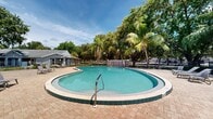 This two-floor property is centered around a generously sized freeform swimming pool that serves as the focal point of the outdoor living area. The pool features a classic kidney-inspired shape with green tile detailing along its edge and includes a convenient entry handrail, making it both functional and visually appealing. Surrounding the pool is a wide, well-maintained brick paver deck that provides ample space for lounging and entertaining. Multiple cushioned lounge chairs and small side tables are arranged throughout the deck, offering comfortable spots to relax and take in the outdoor setting.

The property is richly landscaped with mature tropical vegetation, including tall coconut palms and broad canopy shade trees that frame the space and provide natural privacy. A wrought iron perimeter fence encloses the pool area, maintaining a secure and defined boundary while preserving open sightlines to the surrounding greenery.

Adjacent to the pool, a covered outdoor structure with a low-pitched roof, decorative columns, and French doors provides a shaded gathering area ideal for outdoor dining or socializing. Additional support structures nearby appear to serve utility functions for the grounds. The overall ambiance is distinctly tropical and resort-like, with abundant natural light, lush surroundings, and a relaxed open-air atmosphere that reflects a warm-climate lifestyle.