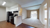 This single-story, one-bedroom, one-bathroom unit offers a functional and efficient layout designed to maximize its compact footprint. The living area is a notable highlight, featuring vaulted ceilings with exposed beams that lend a sense of openness and visual height to the space. Warm neutral tones on the walls complement the plush carpeting throughout the living and bedroom areas, while wood-style flooring anchors the kitchen and entryway zones.

The kitchen is positioned along one wall and opens to the living area via a peninsula-style breakfast bar, creating a seamless connection between cooking and living spaces. White cabinetry provides ample storage, and the kitchen is equipped with black appliances, including a range and refrigerator. A range hood and overhead lighting complete the functional setup.

The living room benefits from a sliding glass door fitted with vertical blinds, offering access to a balcony and allowing natural light to fill the space. A built-in wall unit is also present in the living area.

The bedroom is a well-proportioned, carpeted room with a window that brings in natural light. The bathroom includes a standard tub and shower combination. Overall, the unit presents a clean, neutral palette throughout.