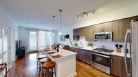 Located at 109 Commerce Dr in Exton, PA, this one-bedroom, one-bathroom residence offers a thoughtfully designed open-concept layout across a single floor. The kitchen and living area flow seamlessly together, anchored by warm hardwood flooring throughout. The kitchen is equipped with full stainless steel appliances — including a gas range, built-in microwave, dishwasher, and a large French-door refrigerator — complemented by rich wood cabinetry, a white tile backsplash, and a peninsula island with pendant lighting overhead, providing additional seating and prep space. The adjoining living room is filled with natural light from large windows and a glass door that opens to an outdoor area, creating an inviting and airy atmosphere. The bedroom features plush carpeting, a generously sized window with sheer curtains, and comfortable proportions suited for a full furniture arrangement. The bathroom is finished with large-format floor tile, a sleek wood-toned vanity with a white countertop, and a glass-enclosed walk-in shower. A dedicated closet provides practical storage. The overall design balances modern finishes with a warm, functional aesthetic throughout.