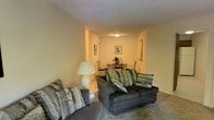 This single-story residence at Mission Viejo Villas offers a straightforward, functional layout designed for comfortable everyday living. The living area is carpeted and furnished with plush, dark upholstered sofas accented by patterned throw pillows, creating a relaxed and inviting atmosphere. Warm, neutral-toned walls run throughout the space, contributing to a cohesive and calm interior palette. A floor lamp provides supplemental ambient lighting, complementing the overhead ceiling fixture that illuminates the room evenly. The living space flows seamlessly into a defined dining area at the rear, where a compact bistro-style table and chairs are arranged beneath a ceiling-mounted light. Framed wall art is displayed throughout, adding simple decorative character to the space. The transition between the living and dining zones is open, enhancing the sense of continuity and depth within the single-floor layout. The overall ambiance is modest and well-maintained, reflecting a practical residential setting suited for individuals or small households seeking a comfortable, move-in ready home within the Mission Viejo Villas community.