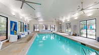 Located at 7305 Village Center Blvd in Fairburn, Georgia, this single-floor indoor pool facility offers a generously sized, climate-controlled aquatic space designed for year-round enjoyment. The centerpiece of the space is a large rectangular swimming pool with crystal-clear turquoise water, featuring clearly marked depth indicators and a surrounding concrete pool deck that provides ample room for movement and relaxation.

The architecture features vaulted ceilings fitted with multiple large industrial-style ceiling fans, ensuring excellent air circulation throughout the space. Abundant natural light streams in through oversized black-framed windows and glass panel garage-style doors positioned along multiple walls, offering views of the surrounding wooded landscape while maintaining a bright and airy atmosphere.

The poolside lounge area is furnished with modern white bucket chairs and cushioned benches accented with blue upholstery, creating a clean, resort-inspired aesthetic. Vibrant, oversized pool-themed wall murals — depicting beach balls, donuts, watermelons, and pool floats — add a playful and energetic character to the space. Posted spa rules and safety signage reflect a well-maintained and safety-conscious environment. The game room designation further underscores the facility's recreational purpose, making it a lively and versatile amenity well-suited for community or group activities.