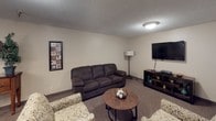 This single-floor space is designed with a practical and comfortable layout, encompassing a dining area and a living room, both finished with neutral beige walls and textured ceilings that lend a consistent, understated character throughout.

The dining area is a well-proportioned room anchored by a substantial dark wood dining table surrounded by matching chairs. A classic chandelier with frosted glass shades hangs overhead, providing warm, focused lighting. Vertical blinds cover a sliding door, allowing natural light to filter into the space while offering privacy. A wall-mounted climate control unit and baseboard heating ensure year-round comfort. Decorative accents, including a framed wall piece and a small console table, add character to the room.

The living room is a relaxed, open space centered around a large, dark upholstered sofa complemented by patterned accent chairs. A round wooden coffee table sits at the center, while a wall-mounted flat-screen television and media console provide an entertainment focal point. Soft carpeting underfoot and a textured ceiling contribute to the room's cozy, welcoming ambiance, making it a practical space for everyday living.