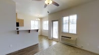 This single-floor space features an open-concept layout that seamlessly connects the living and dining areas with the kitchen. The living area is finished with hardwood-style flooring that transitions to carpet, adding warmth and texture underfoot. Neutral-toned walls throughout create a clean, versatile backdrop. A ceiling fan with a built-in light fixture overhead provides both comfort and ambient lighting, while a chandelier in the adjacent dining area adds a decorative touch.

The kitchen is partially separated by a raised breakfast bar with a wood-trimmed counter overhang, offering a casual seating option and a natural visual divide between spaces. Multiple windows allow generous natural light to fill the interior, enhancing the airy feel of the open layout. A glass-paneled door provides direct access to an outdoor balcony, extending the living space and offering outdoor views.

A wall-mounted HVAC unit ensures year-round climate control. The overall ambiance is bright, clean, and functional, making it a practical and comfortable single-level living space.