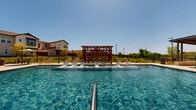 Located at 13000 Saginaw Blvd in Fort Worth, Texas, this community outdoor amenity area offers a well-appointed, resort-inspired gathering space designed for relaxation and recreation. At the heart of the space is a generously sized, crystal-clear swimming pool with a smooth concrete surround lined with lounge chairs, ideal for soaking up the Texas sun. A striking wood pergola serves as a central architectural focal point beside the pool, providing a shaded seating area with an open, airy design that complements the surrounding landscape. On the opposite side of the pool, a covered pavilion with a standing-seam metal roof houses an outdoor kitchen complete with a built-in grill and fireplace feature, making it a functional hub for outdoor entertaining. The pool area transitions seamlessly into a broad, well-maintained open lawn that offers ample space for recreation and community gatherings. Ornamental landscaping with native grasses and shrubs lines the perimeter, adding texture and color throughout the grounds. The entire area is enclosed by a black iron fence, providing security while maintaining open sightlines to the surrounding neighborhood. Under the clear blue Texas sky, this amenity space delivers a vibrant and inviting outdoor experience for the community.