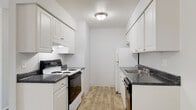 This single-floor unit offers a clean, functional layout with a consistent neutral color palette throughout. The galley-style kitchen is efficiently designed with white shaker cabinetry lining both walls, dark speckled countertops, and a full suite of appliances including an electric range, refrigerator, and dishwasher. A stainless steel sink and under-cabinet range hood add practical utility, while wood-look plank flooring ties the space together with a warm tone.

The living area is generously proportioned, featuring plush wall-to-wall carpeting, a baseboard heating unit, and a large sliding door dressed with vertical blinds that allows natural light to fill the room. A ceiling fan with a light fixture is present in a secondary area near the entryway, which also benefits from wood-look flooring and built-in closet storage.

The bedroom is carpeted and offers a roomy feel, complemented by a double-door closet providing ample storage. The full bathroom maintains the space's clean aesthetic with an all-white finish, a tub-shower combination with tiled surround, a single vanity with a large mirror, and wood-look flooring. Overall, the unit presents a bright, move-in-ready atmosphere with practical finishes throughout.